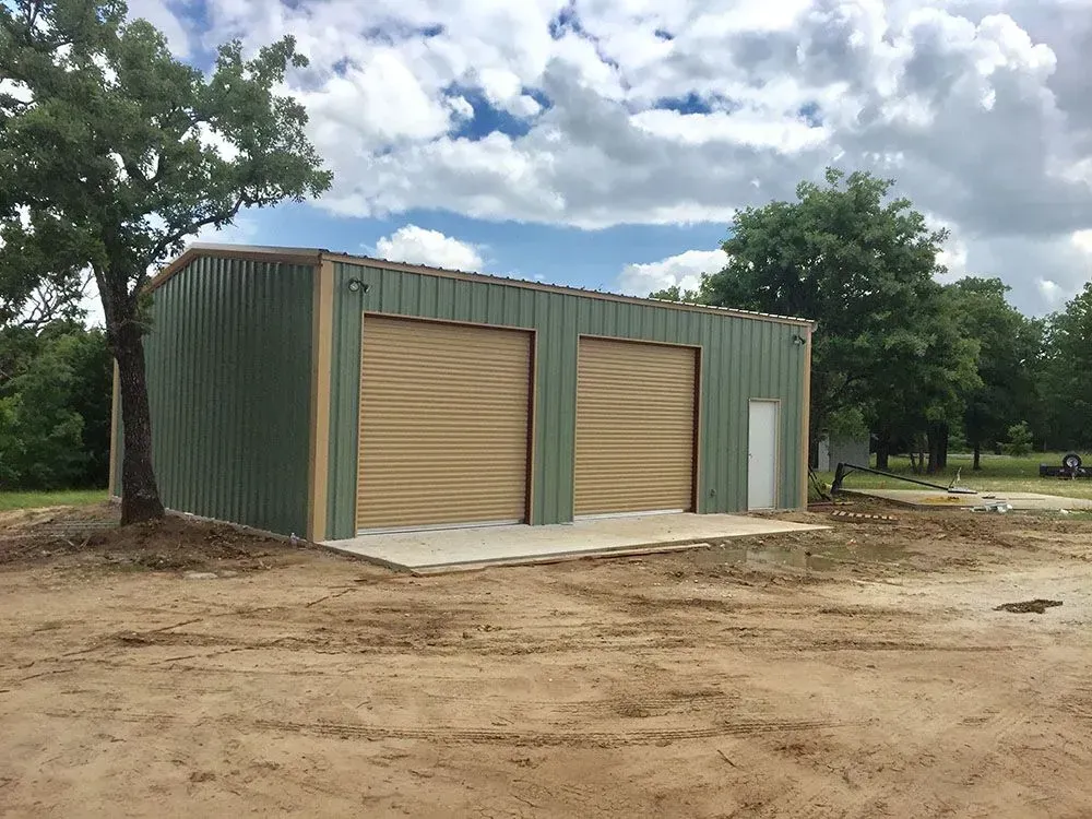 A green and tan metal workshop building with two roll-up doors and a side door, situated on a dirt lot with trees.