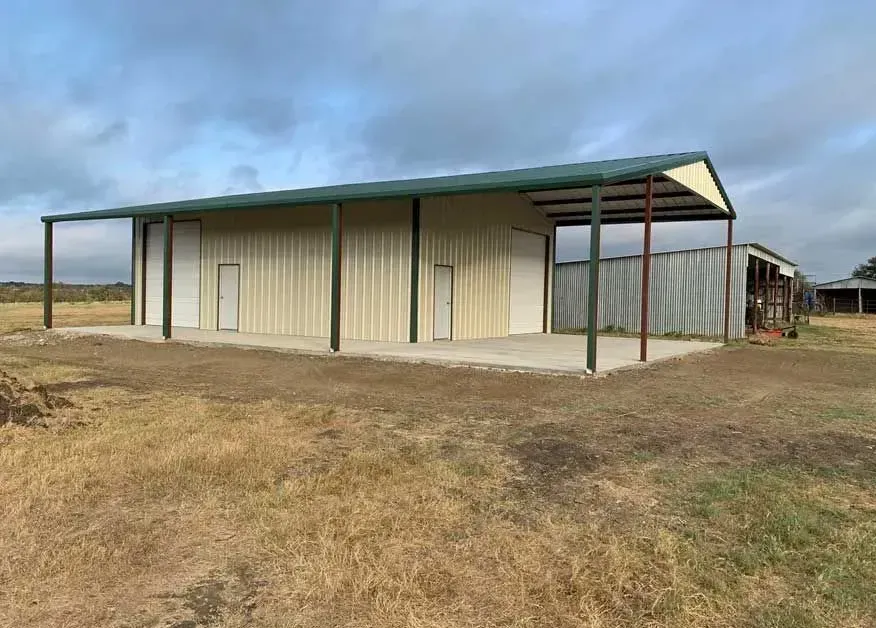 A tan metal building with a dark green roof and a covered porch, set on a flat, grassy field under a cloudy sky.