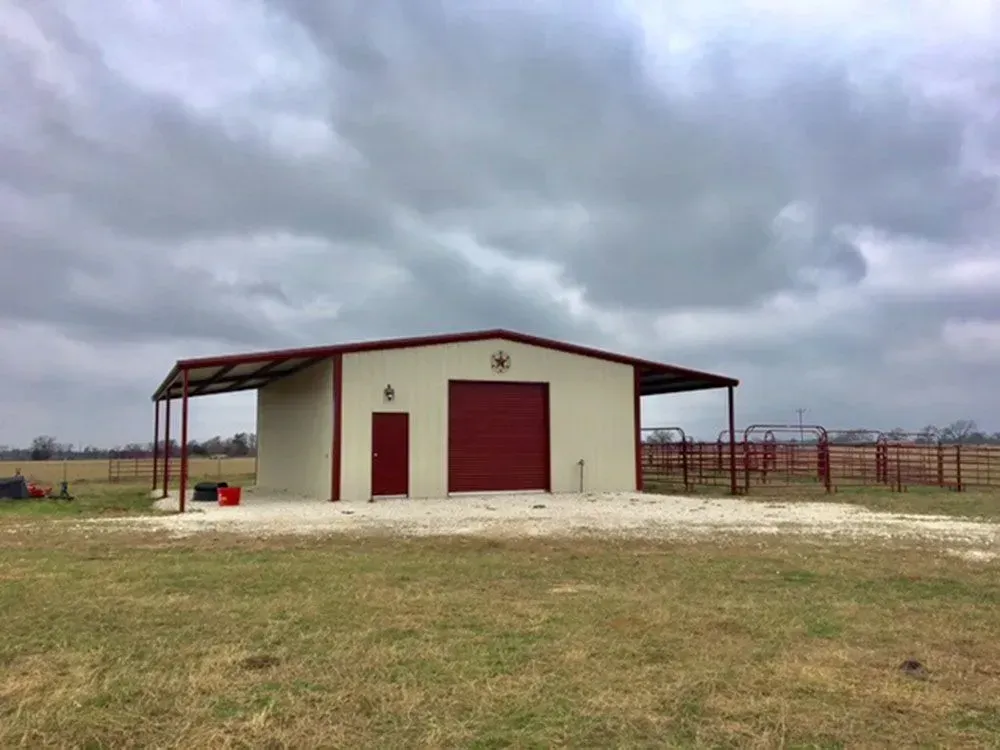 A tan metal workshop with a red door and roll-up garage door sits in a field under a cloudy, overcast sky.