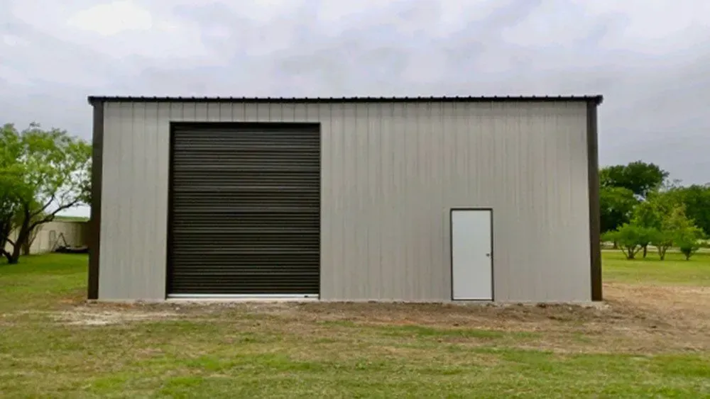 A grey metal storage building with a large dark rolling door and a single white entry door, set in a grassy field.