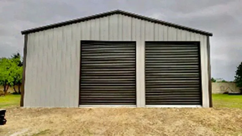 A light-colored metal storage building with two dark-colored rolling garage doors, situated on a grassy lot.