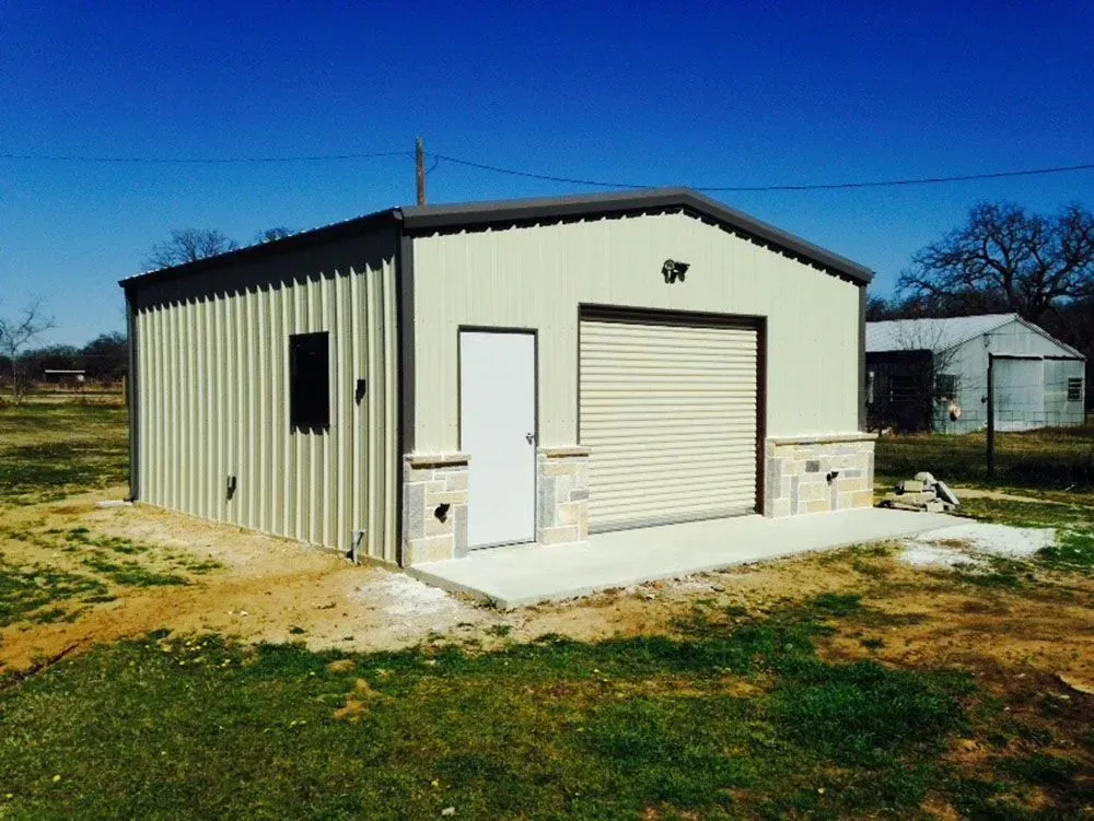 A modern beige metal garage with a roll-up door, a white side door, and stone accents, set on a rural property.