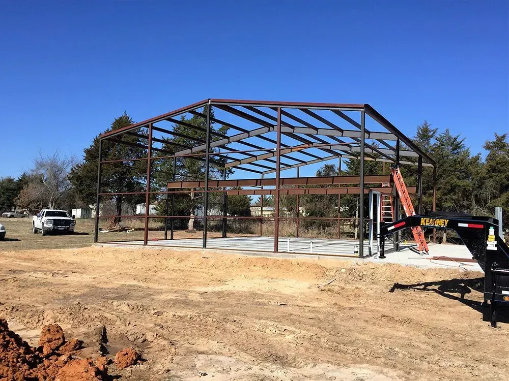 The steel frame of a building under construction, set on a concrete slab in an open, grassy area under a blue sky.