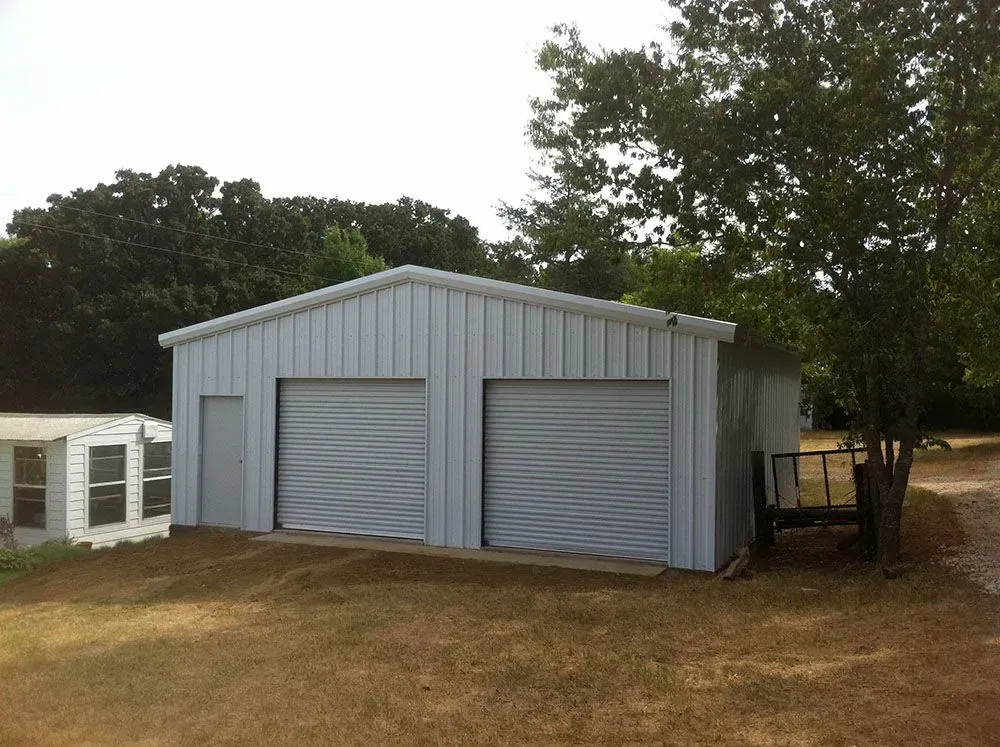 A light-colored metal garage with two roll-up doors and a side entrance, situated on a grassy lot near some trees.