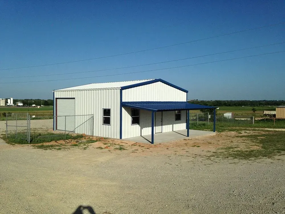 A white metal storage building with blue trim and a covered front porch, situated on a gravel lot under a clear sky.
