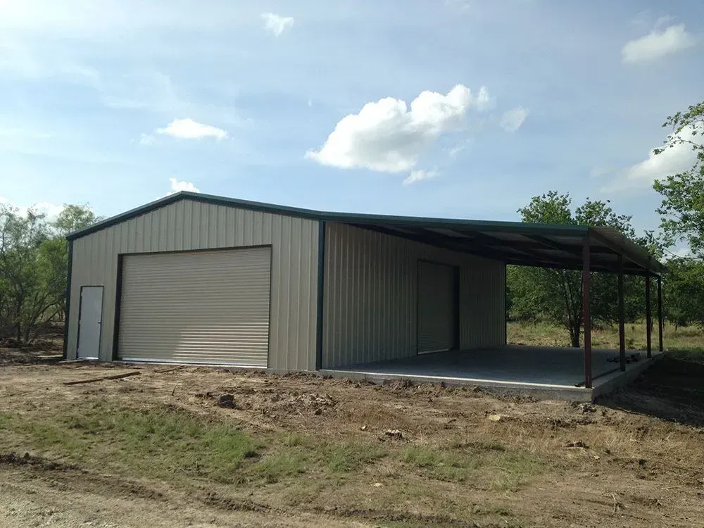 A beige metal building with a garage door, side entry door, and an attached open carport on a dirt lot under a blue sky.