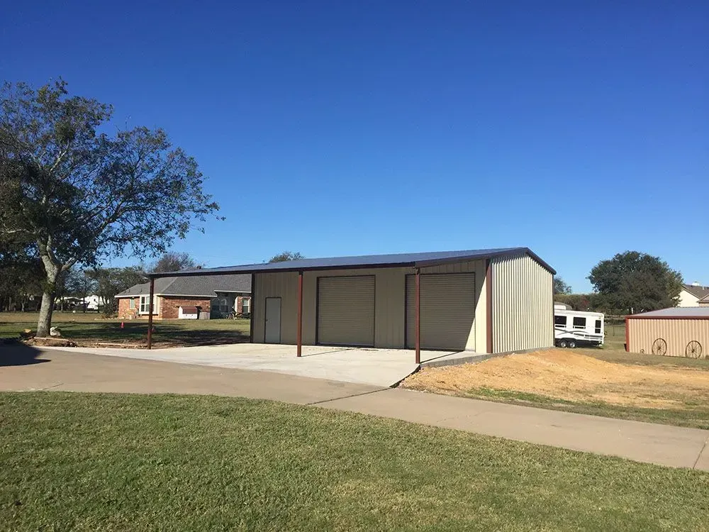 A metal storage building with two garage doors and an open-sided carport area next to a paved driveway on a sunny day.