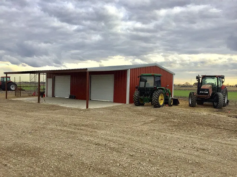 A red metal shed with a covered porch and two tractors parked on a gravel lot under a cloudy sky.