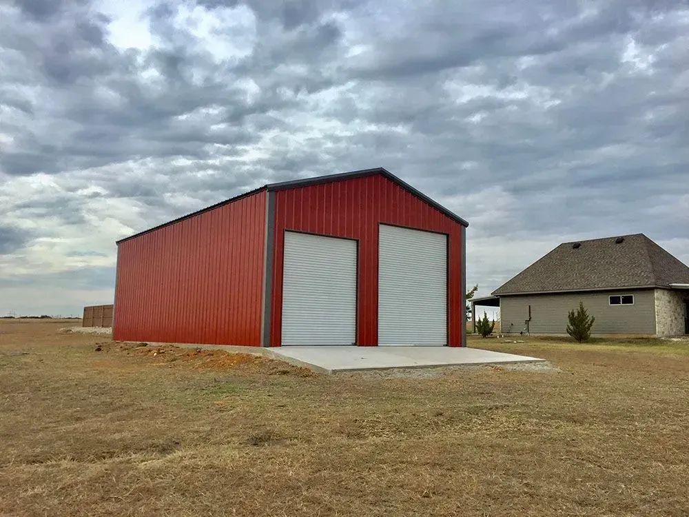 A red metal garage building with two roll-up doors stands in a grassy field next to a house under a cloudy sky.