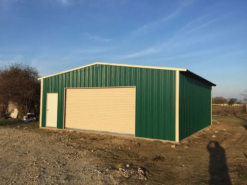 A green metal garage building with a beige roll-up door and a single side door, set on a gravel lot under a clear sky.