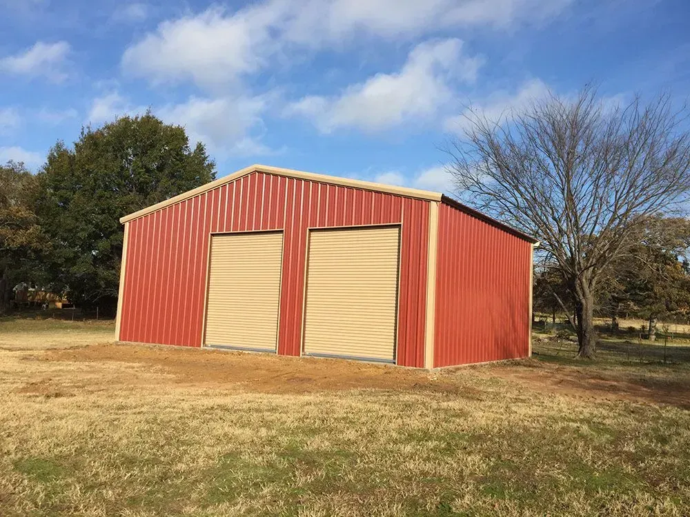 A red metal barn with two tan roll-up garage doors stands in a grassy field under a blue sky with scattered clouds.