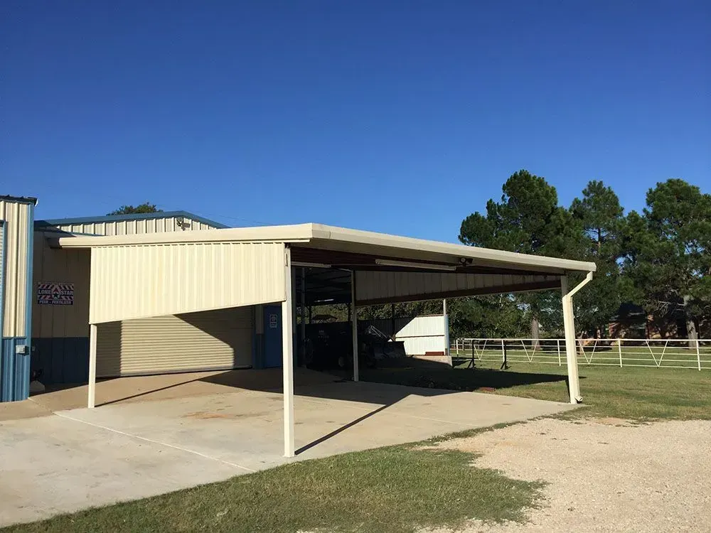 An outdoor carport with a sloped, beige metal roof and matching side panel, attached to a building on a rural property.