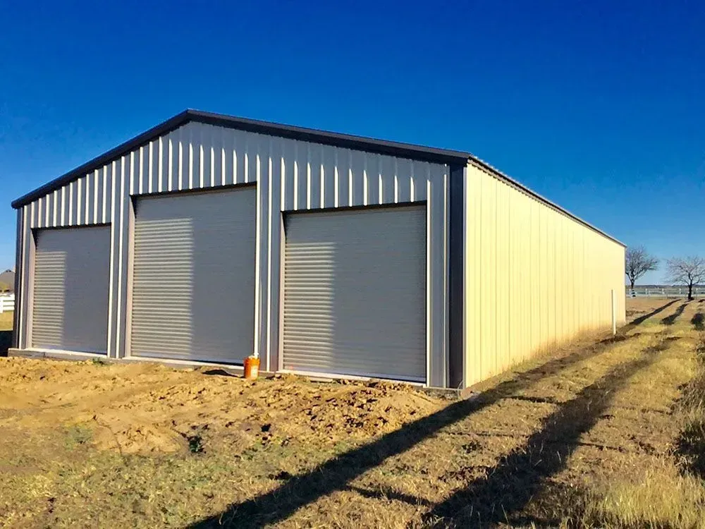 A rectangular, tan metal building with three roll-up doors, set on a dirt lot under a clear blue sky.