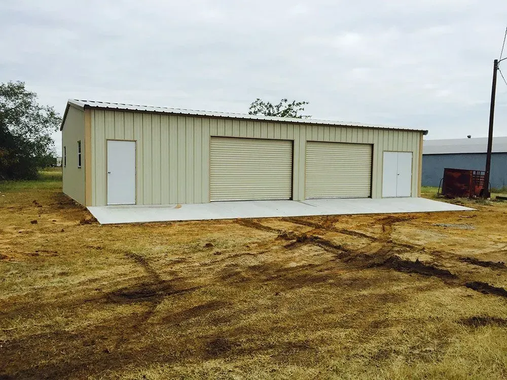 A light-colored metal workshop building with two garage doors and two entry doors, set on a grassy lot.