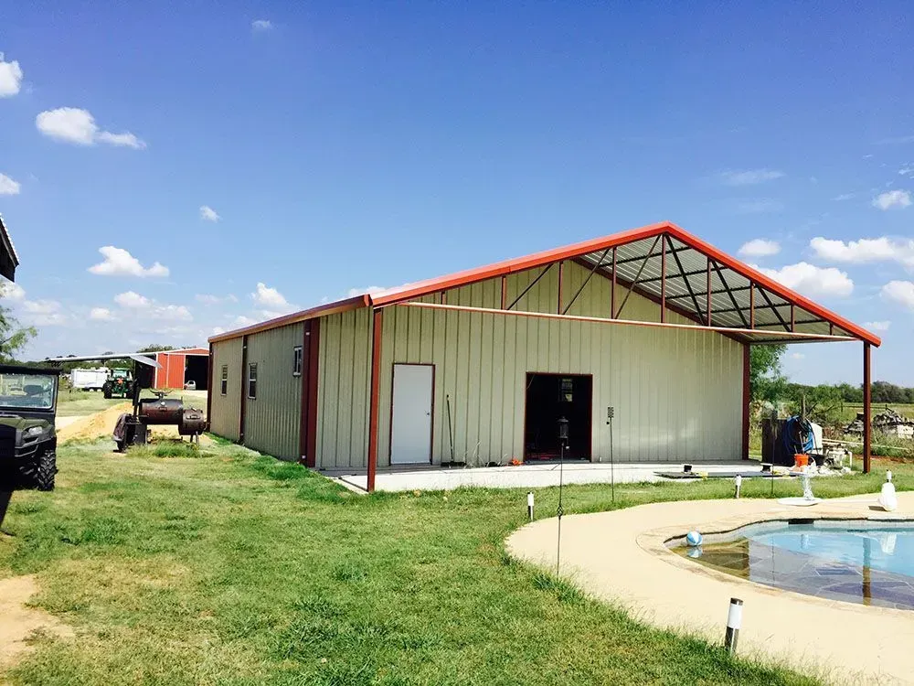 A tan metal workshop building with a red-trimmed roof stands next to a backyard swimming pool under a sunny sky.