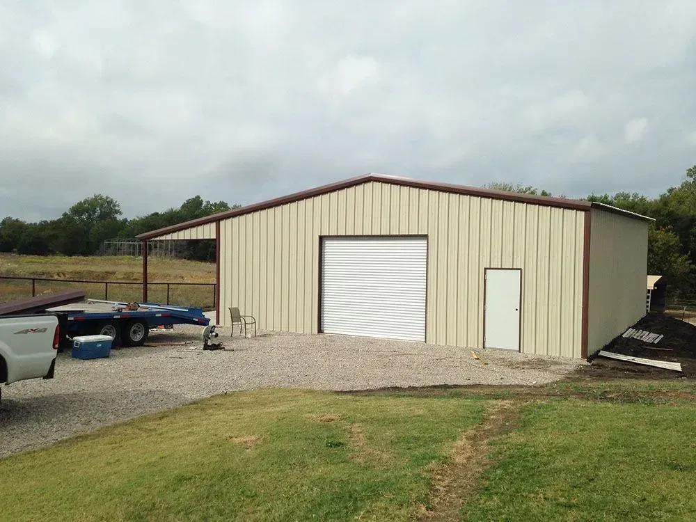 A tan metal shop building with a roll-up door and side entry door, situated on a gravel lot near a grassy field.