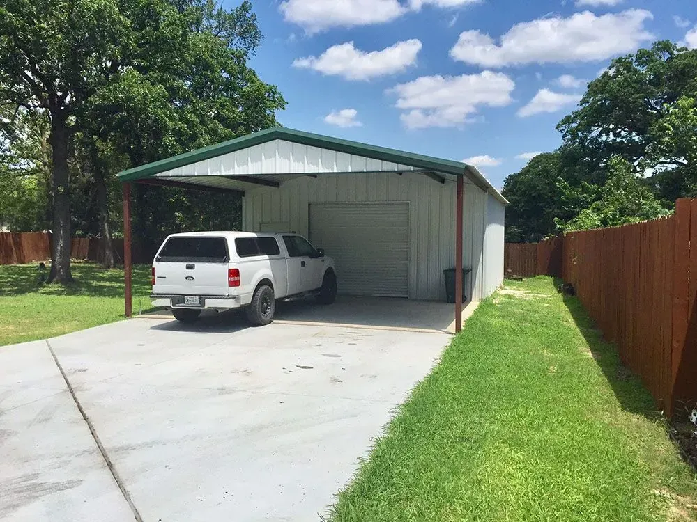 A white pickup truck with a camper shell parked under a metal carport next to a green-roofed shed on a sunny day.