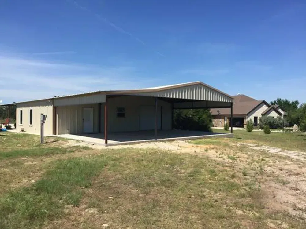 A tan metal building with an attached carport sitting on a gravel lot under a bright blue sky, with a house nearby.
