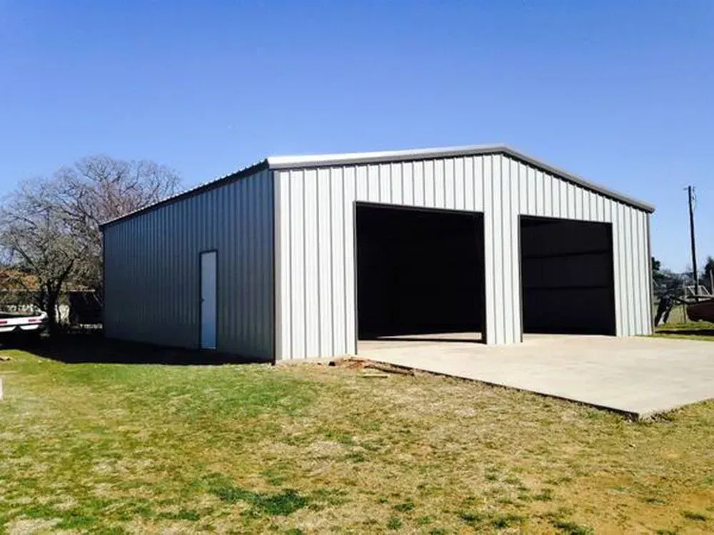 A light-gray metal garage with two wide open doorways and a single side entry door sits on a concrete pad in a yard.