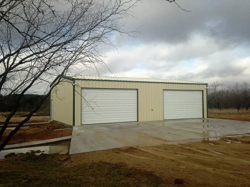 A tan metal garage with two white roll-up doors, set on a concrete slab under a cloudy sky near bare trees.