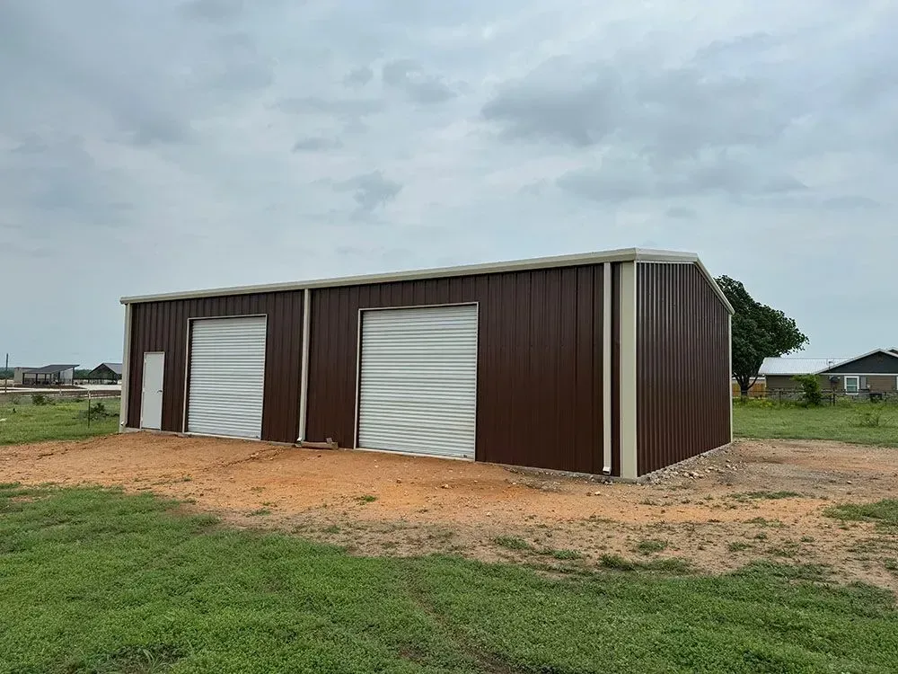 A brown metal shop building with two white garage doors and a side entry door, set on a grassy lot under a cloudy sky.