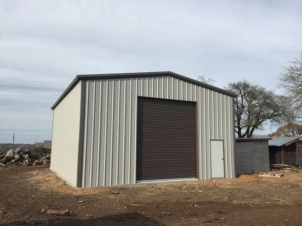 A light-colored metal workshop building with a dark brown roll-up door and a single side door on a dirt lot.