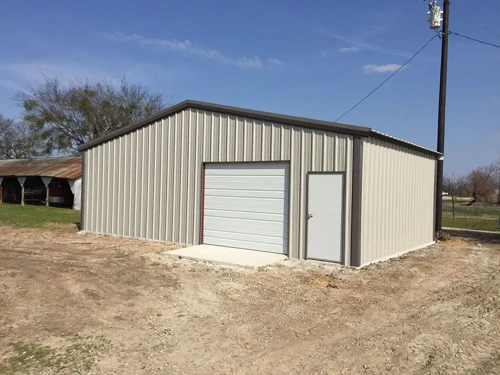 A tan metal workshop with a white roll-up garage door and a side entry door, located on a dirt lot under a blue sky.