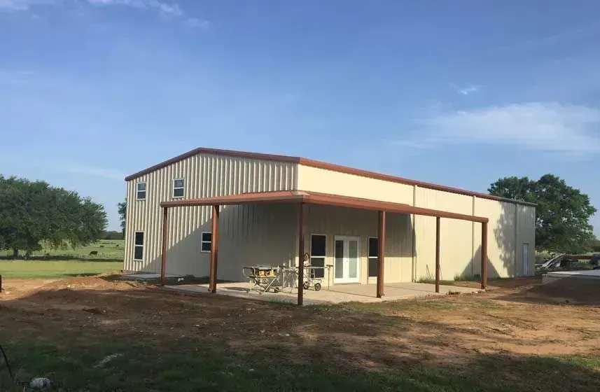 A large, beige metal building with a covered patio area set against a rural landscape with trees and a blue sky.