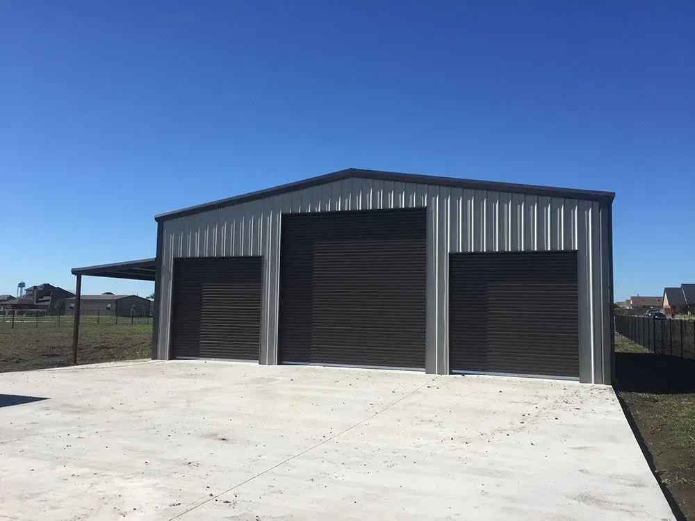 A modern metal barn with three dark garage doors and an attached side porch on a concrete slab under a clear blue sky.