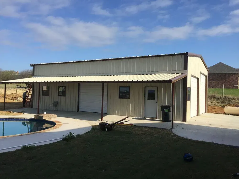 A beige metal workshop with a covered porch and garage doors next to a backyard swimming pool under a blue sky.