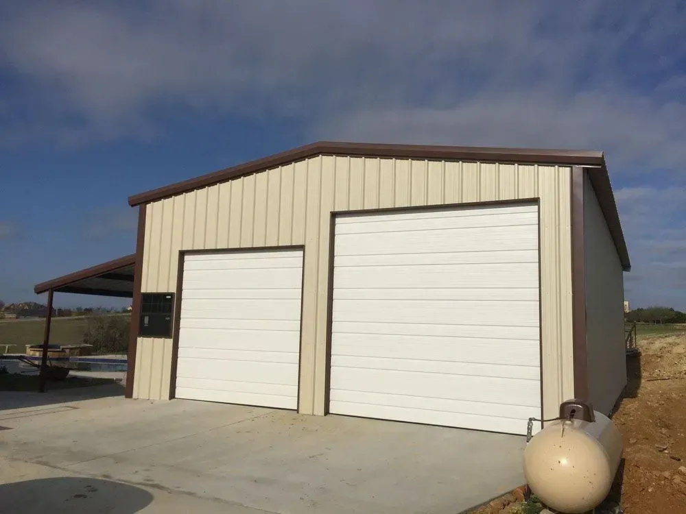 A light-colored metal workshop building with two garage doors and an attached side porch under a blue sky.