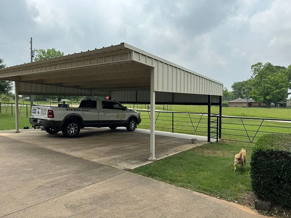 A white pickup truck parked under a metal carport on a sunny, grassy property with a small dog standing on the lawn.