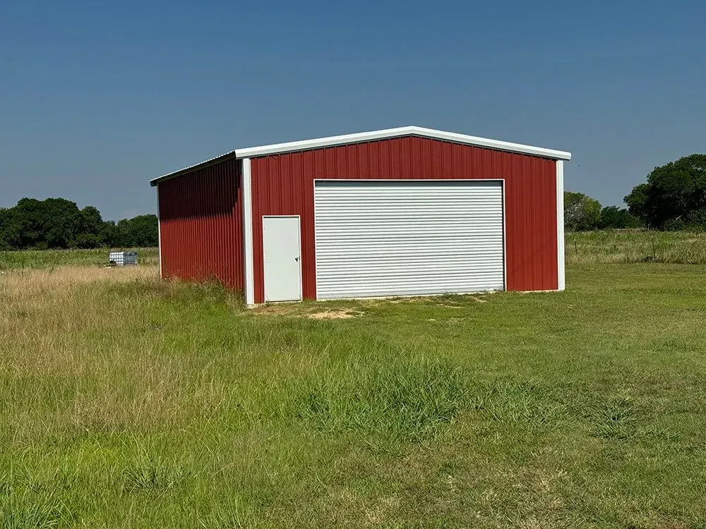 A red metal barn with a white door and a large white overhead garage door set in a grassy field under a clear blue sky.