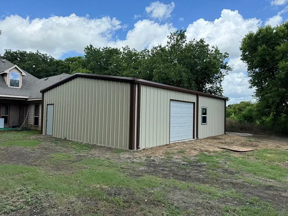 A beige metal garage with a white roll-up door and a side entry door, standing next to a house on a grassy lot.