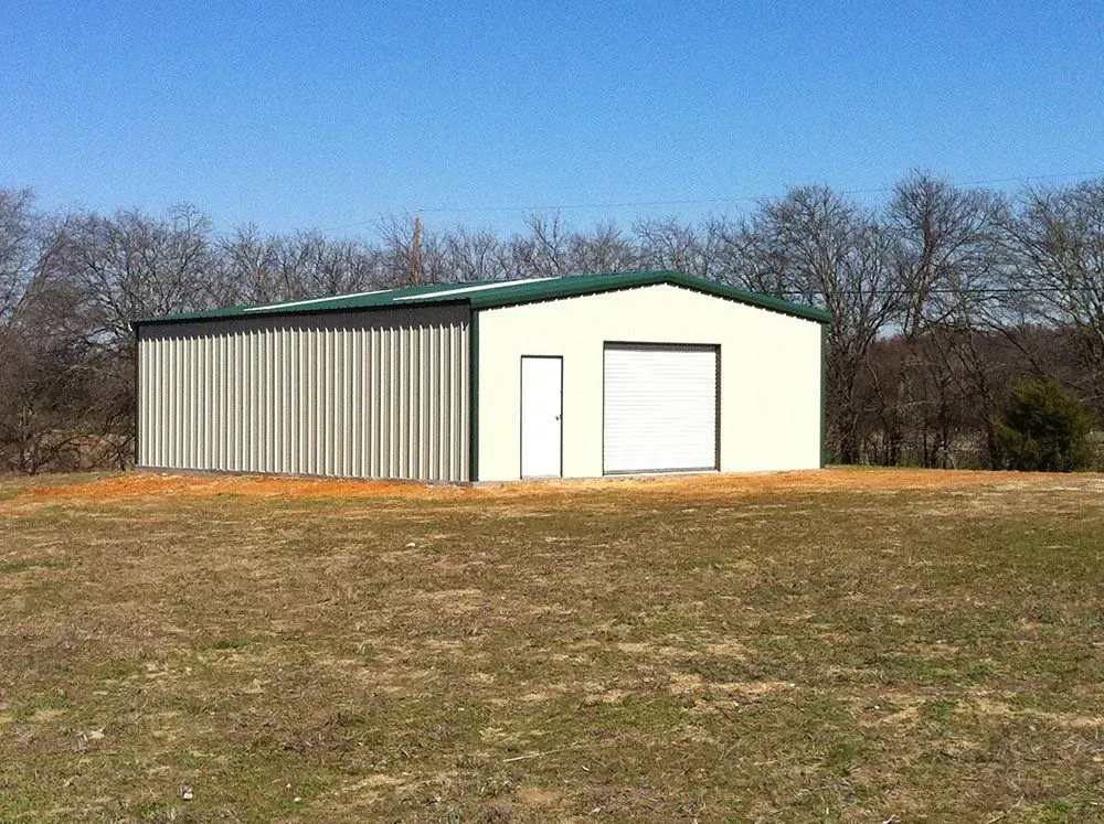 A rectangular metal warehouse building with off-white walls, a green roof, and a single roll-up door in a grassy field.