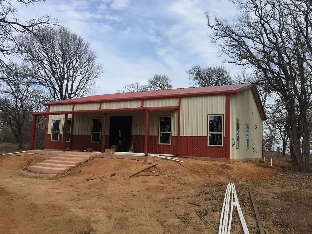 A newly constructed metal building with tan and red siding, a red roof, and a porch, surrounded by trees on a dirt lot.