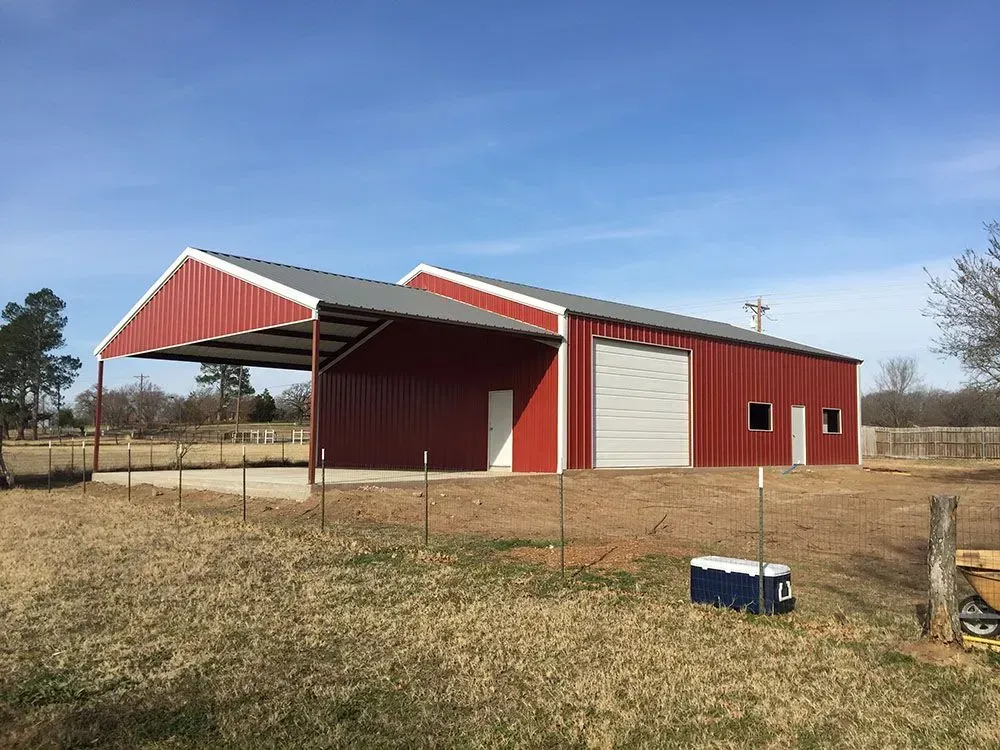 A red metal building with a white garage door and an attached open-air carport in a grassy field under a blue sky.