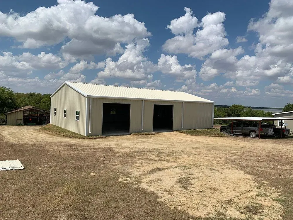 A light-colored metal workshop building with two garage doors sits on a dirt lot under a blue sky with scattered clouds.
