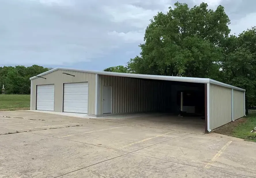 A tan metal garage building with two white roll-up doors and an adjacent open-sided carport on a concrete driveway.