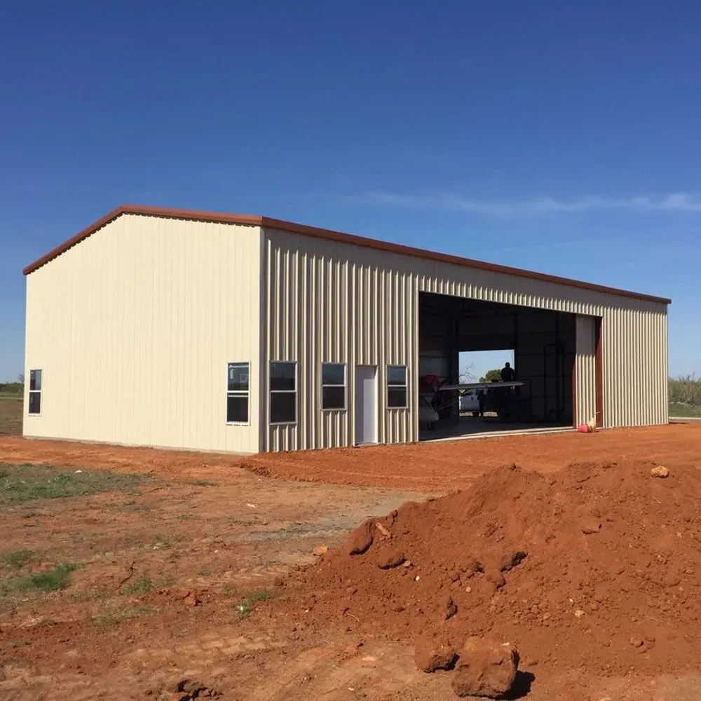 A beige metal-sided building with brown trim and an open garage bay sits on red dirt under a clear blue sky.