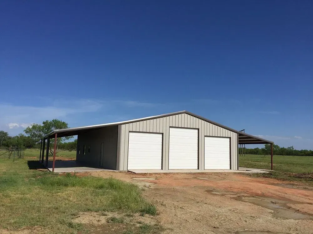 A metal workshop building with three white garage doors and side lean-to structures, situated in a grassy field.