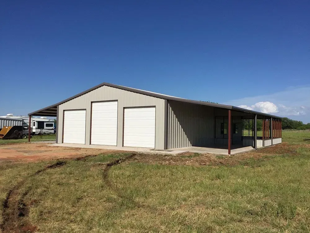 A beige metal building with three white roll-up garage doors stands in a grassy field under a clear blue sky.
