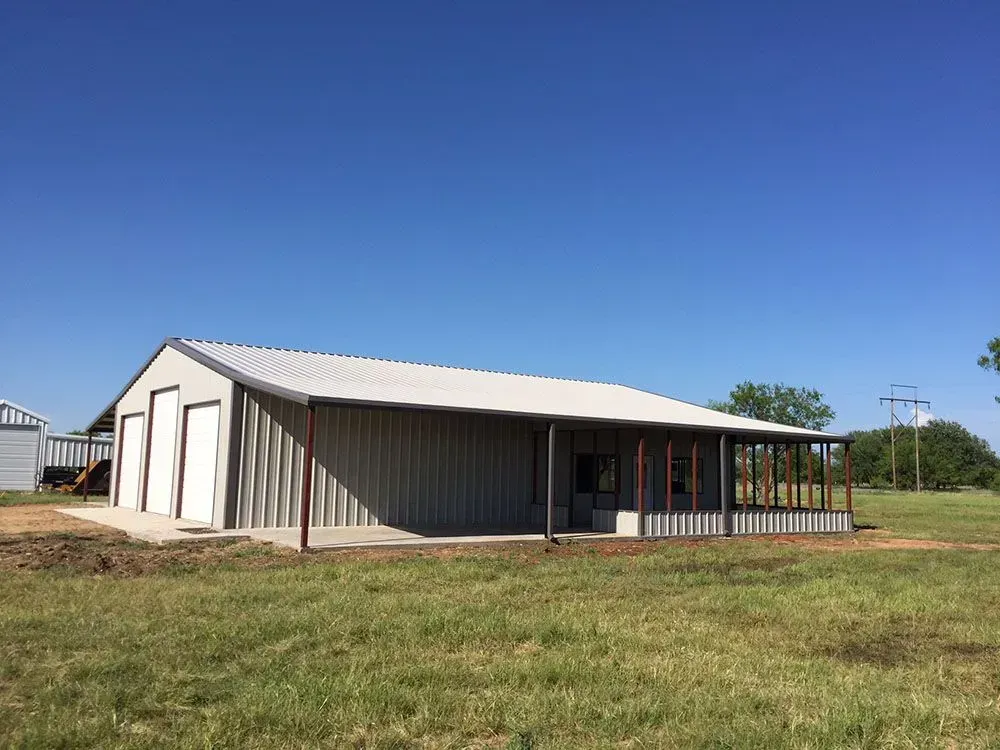 A light-colored metal barn with garage doors and a side porch set in a grassy, open field under a clear blue sky.