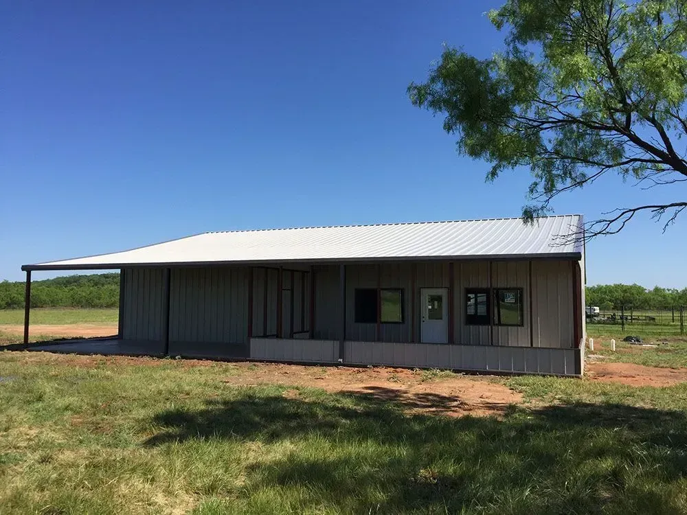 A metal barndominium-style building with a white roof and tan siding sits in a sunny, grassy field under a blue sky.