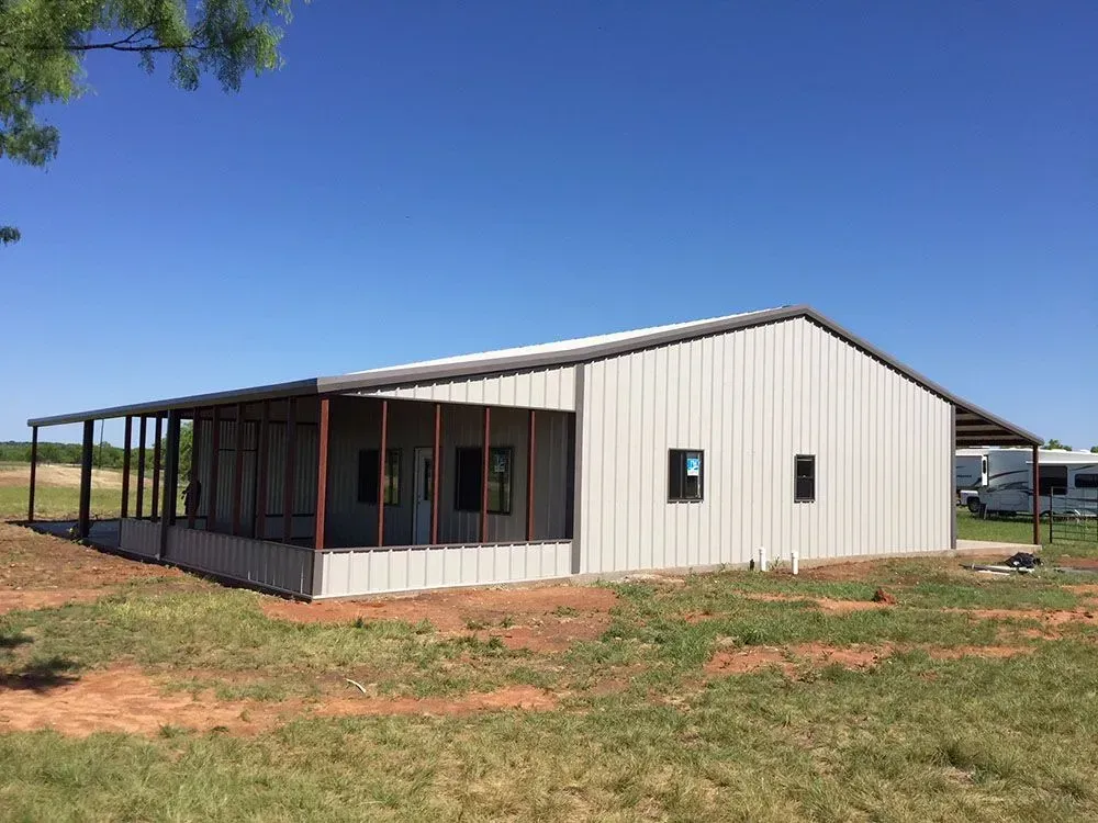 A modern light-gray metal barndominium with a covered porch set against a clear blue sky.
