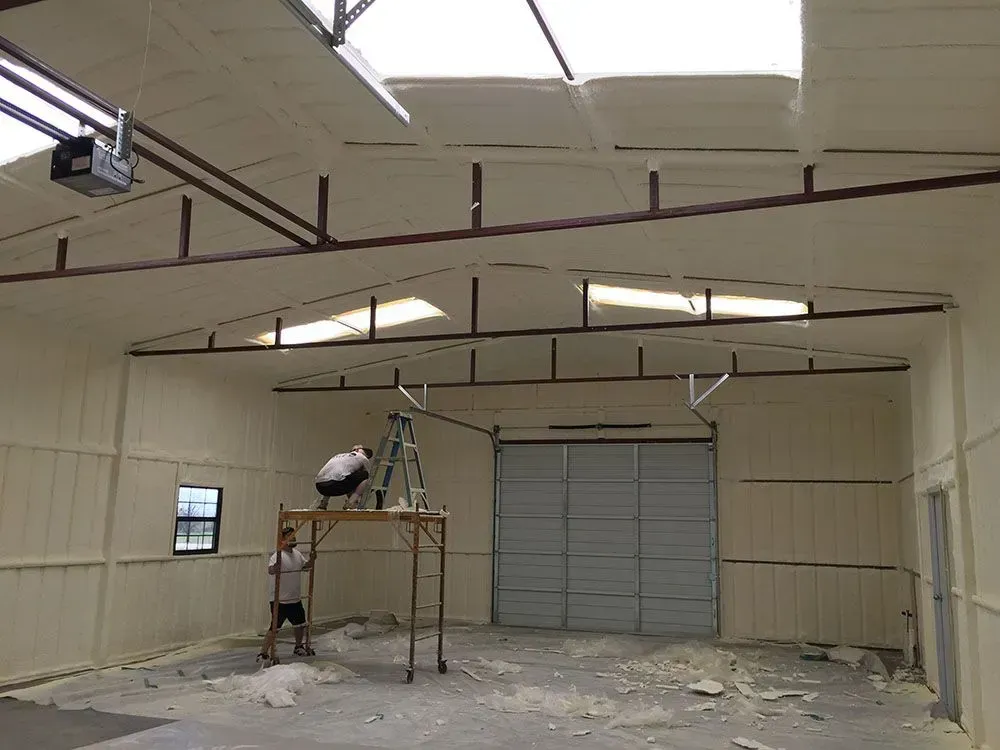 Two people work on scaffolding in a large warehouse space with off-white spray-foam insulation on the walls and ceiling.