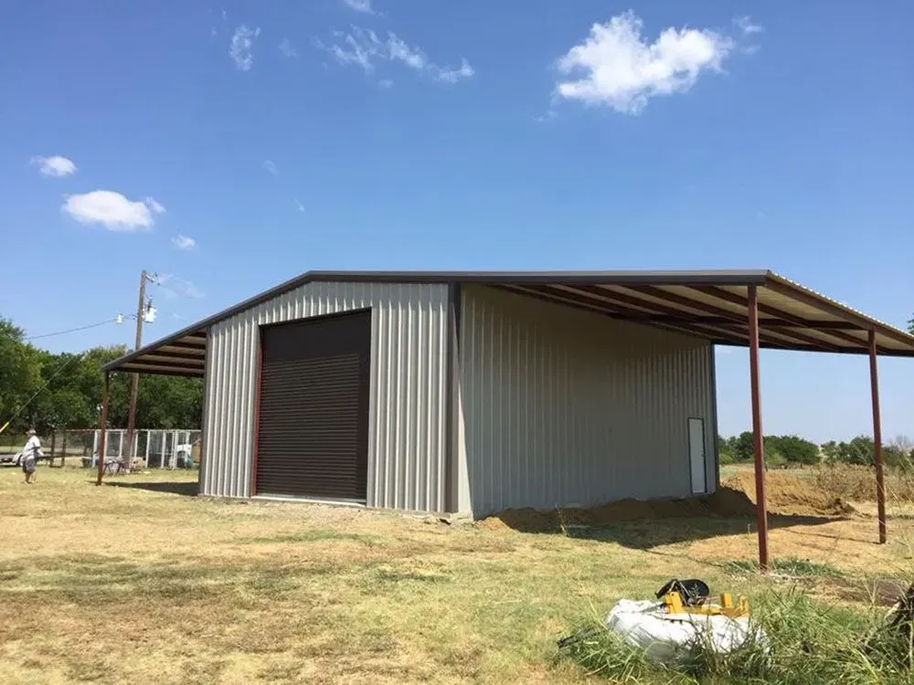 A beige metal workshop building with a large roll-up door and a side lean-to shelter under a clear blue sky.