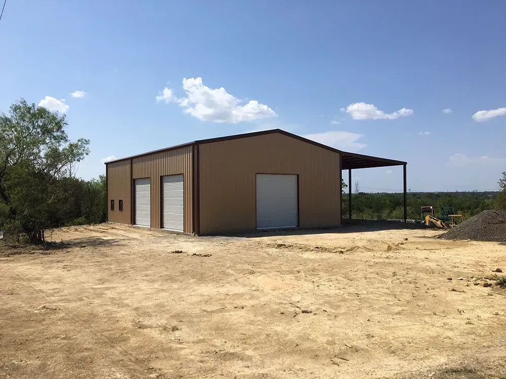 A tan metal warehouse with several roll-up garage doors sits on a dirt lot under a bright blue sky.