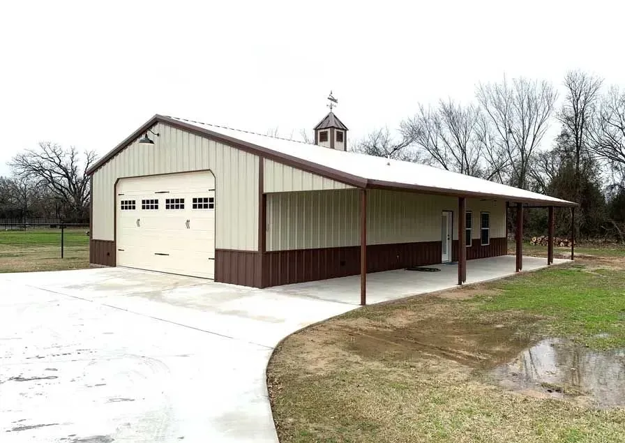 A tan and brown metal building with a garage door, a covered porch, and a cupola in a rural setting.