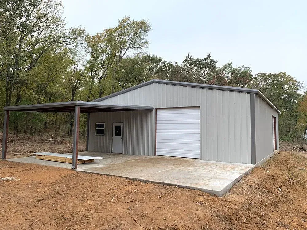 A gray metal workshop with a white roll-up door, a side entry door, and an attached covered concrete patio in a woods.